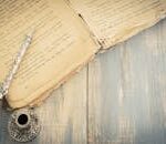 Close-up of an antique book with an ornate quill and ink pot on a wooden surface, evoking a classic vintage feel.